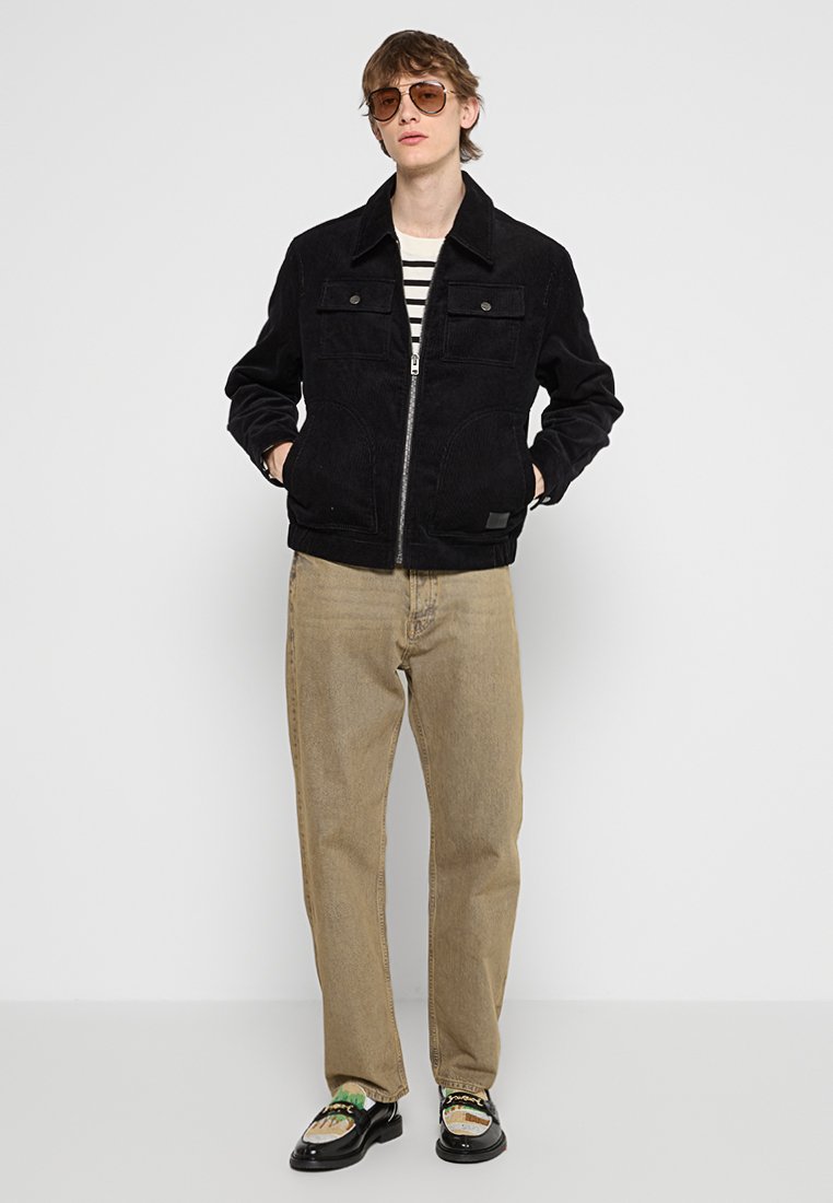 Veste en velours côtelé noir avec fermeture éclair et deux poches avant. Portée par-dessus une chemise rayée, associée à un pantalon beige et des chaussures noires.