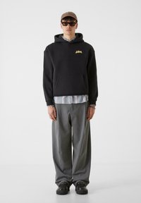 Črna kapuca z zlatim logotipom, prek modro-belega črtastega srajca. Sive široke hlače in črne čevlje. Model nosi sončna očala in kapo.