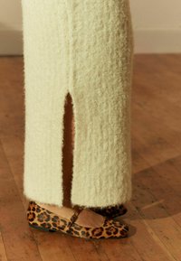 Cremefarbene Strickskirt mit vorderer Schlitz, kombiniert mit Ballettschuhen im Leopardenmuster, die einen dünnen Riemen und eine flache Sohle haben, auf einem Holzboden.
