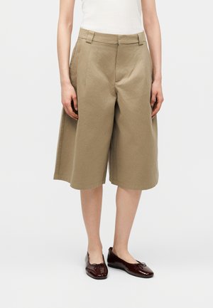 Person iført knælang, vid buks i khakifarve, hvid top og mørkebrune flade sko, stående mod en ensfarvet baggrund.