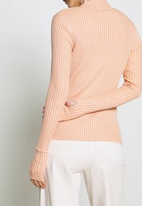 Maglione a collo alto a coste in morbido tessuto pesca, con maniche lunghe, design aderente e aperture per il pollice, abbinato a pantaloni bianchi a vita alta.