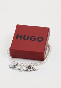 Perlo apyrankė su sidabriniu žavesiu "Lucky You" šalia raudonos dėžutės, ant kurios juodu raštu užrašyta "HUGO" ant balto fono.