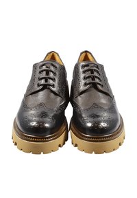 Scarpe in pelle nera con dettagli brogue a punta, caratterizzate da una suola in gomma spessa e testurizzata di colore beige. I lacci sono ben visibili.