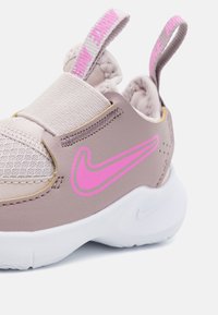 Vaaleanruskeat tennarit, joissa on verkkomateriaali, joustava hihna, vaaleanpunainen Nike-logo, pyöreä valkoinen pohja ja vetotappi kannassa.