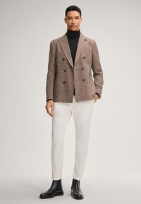 Bruine geruite dubbelgebreide blazer met een gestructureerde afwerking, gecombineerd met een zwarte coltrui, crèmekleurige broek en zwarte enkellaarsjes.