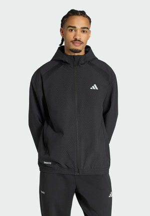 Man met gevlochten haar, die een zwarte, gestructureerde Adidas-jas met capuchon en bijpassende zwarte broek draagt, staand tegen een effen achtergrond.