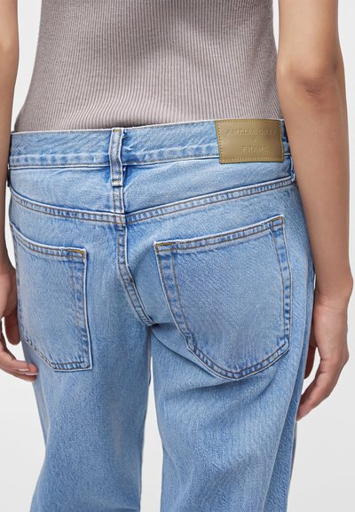 Vue de dos d'une personne portant un jean en denim bleu clair avec deux poches arrière et une étiquette de marque en cuir beige sur la taille.