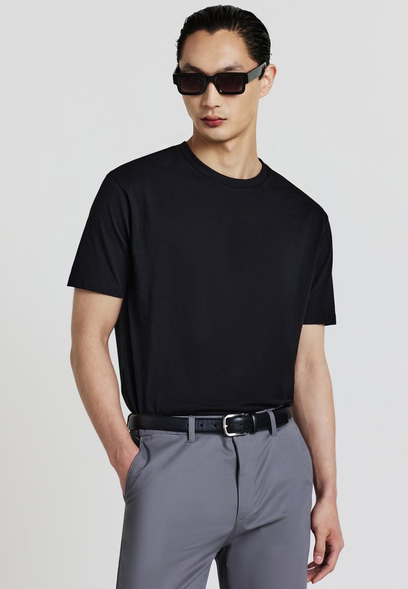 Uomo con occhiali da sole neri, maglietta nera, pantaloni grigi e cintura nera, in posa con una mano in tasca su uno sfondo bianco.