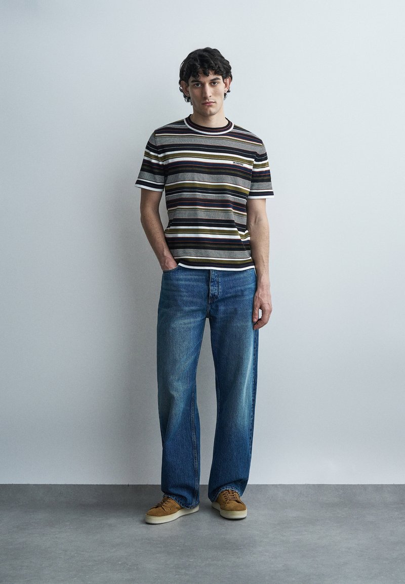 Young man standing with hands in pockets wearing a multicolored striped t-shirt, wide-leg blue jeans, and brown sneakers against a plain wall.