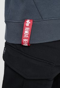 Felpa grigia con polsini a coste, caratterizzata da un'etichetta rossa con testo bianco. Pantaloni cargo neri con dettagli visibili delle tasche e cuciture.