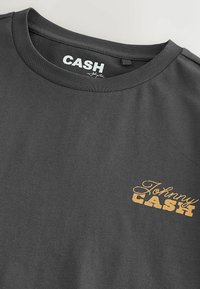 Sötétszürke rövid ujjú póló kerek nyakkivágással és a bal mellrészen sárga "Johnny Cash" logóval nyomtatva.