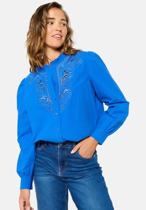 Blouse bleue avec un design floral brodé, devant à boutons, et manches longues bouffantes, associée à un jean en denim taille haute.