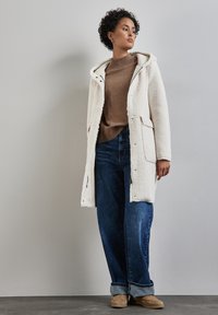 Crème-kleurige fleece jas met een capuchon, knoopsluiting en zijzakken, gedragen over een bruine trui en blauwe wijde jeans.