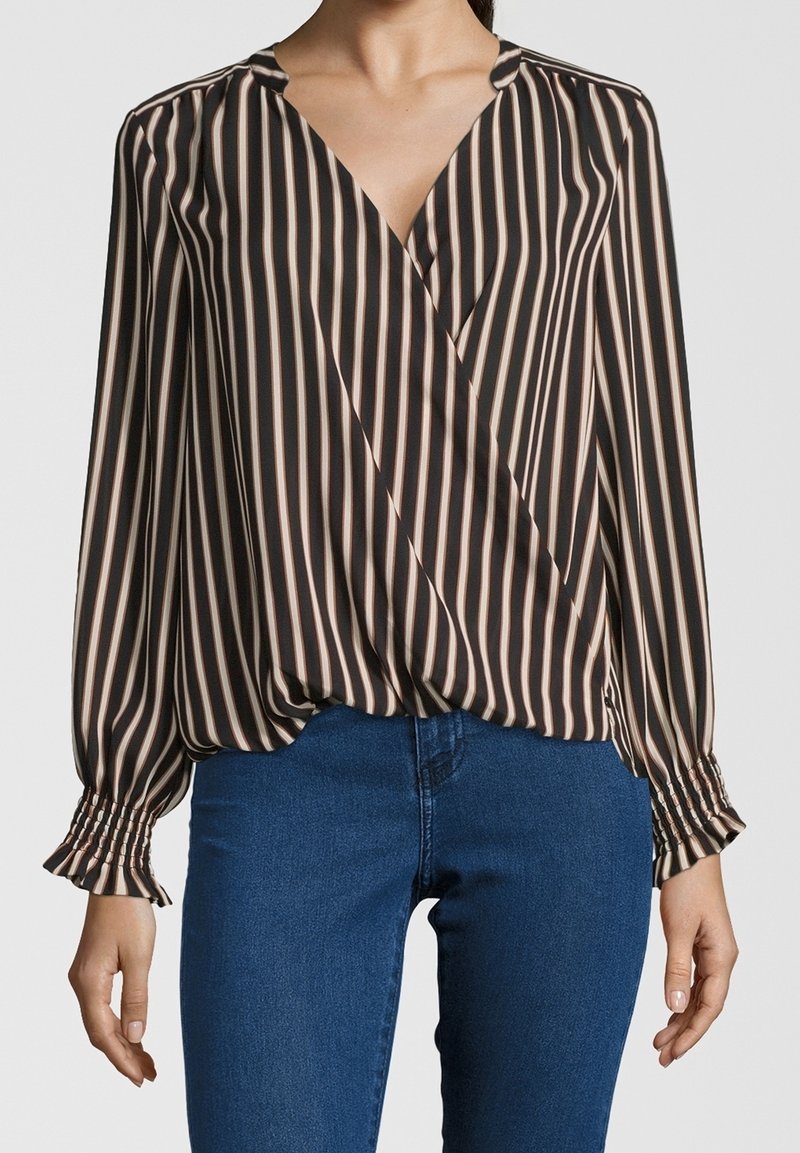 Femme portant un chemisier noir à rayures beiges verticales et manches longues, associé à un jean bleu, montrée du cou aux hanches.