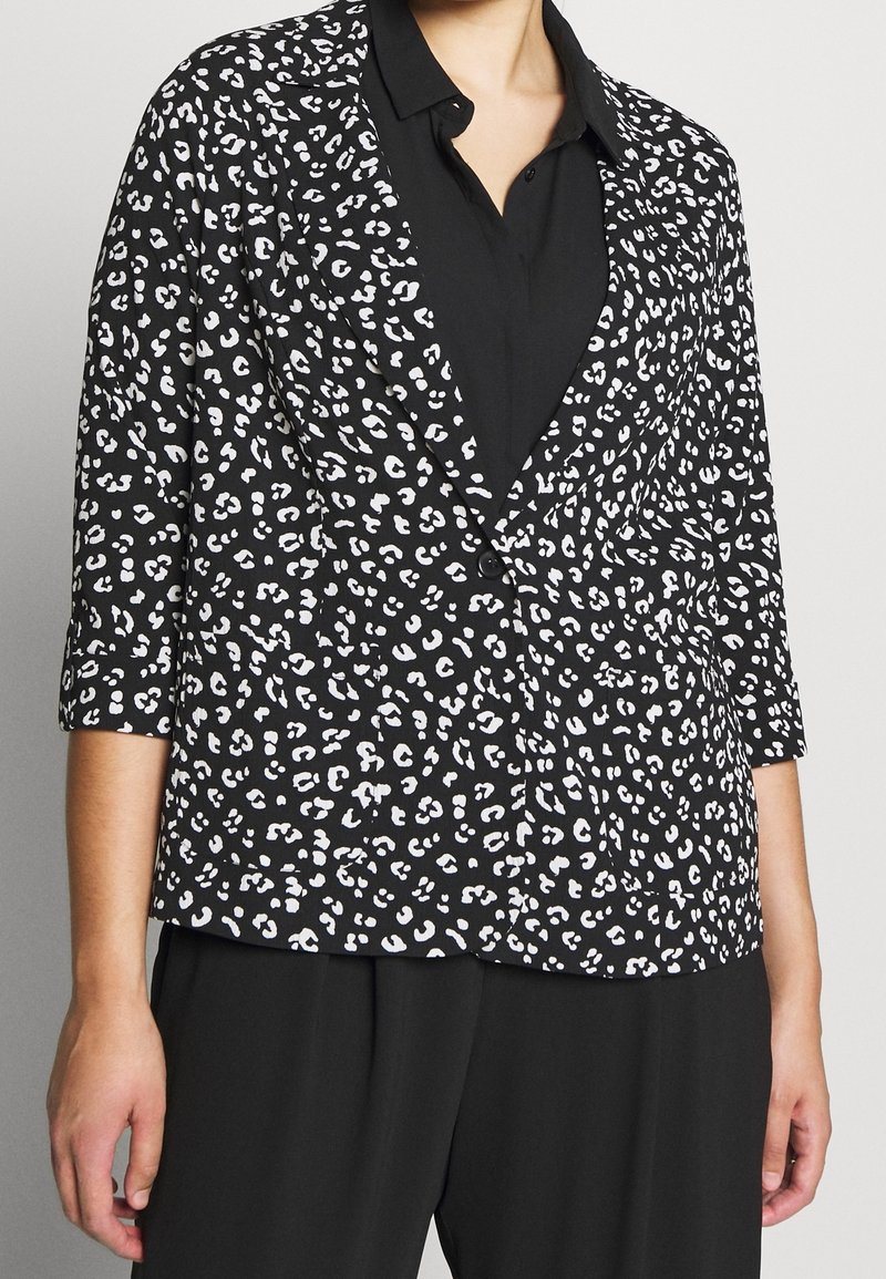 Personne portant un blazer noir avec imprimé léopard blanc, manches trois-quarts, et une chemise noire boutonnée en dessous.