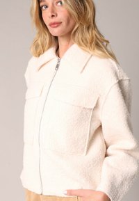 Witte fleece jas met een rits aan de voorkant, grote borstzakken en een kraag. Het textuurmateriaal voegt diepte toe aan het zachte ontwerp.