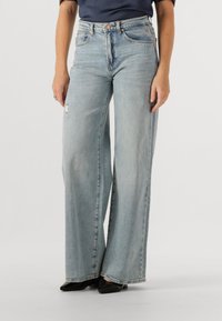 Lichtblauwe wijde jeans met een hoge taille, gemaakt van denim, met een versleten detail op de linker bovenbeen. Zwarte puntige schoenen worden gedragen.