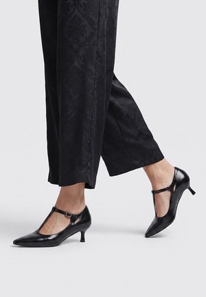 Piernas con pantalones negros estampados recortados y zapatos negros de tacón kitten con tira en T sobre un fondo gris claro.