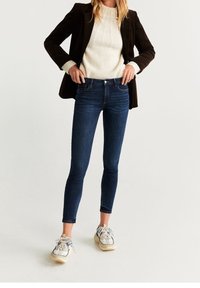 Jeans ajustados azules de cintura media, combinados con un suéter crema de punto trenzado y un blazer oscuro, complementados con zapatillas de deporte estampadas.
