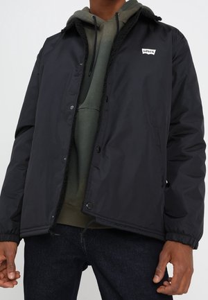 Homme portant une veste noire Levi's par-dessus un sweat à capuche vert olive et un jean foncé, debout les mains détendues le long du corps.