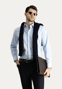 Camisa de botón azul claro, suéter de punto marino sobre los hombros, pantalones oscuros, cinturón marrón y un portafolio marrón texturizado con un acabado suave.