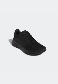 Zapatilla deportiva negra con parte superior de malla, sistema de cordones y tres franjas como acentos en el costado. Suela de goma gruesa para tracción y soporte.