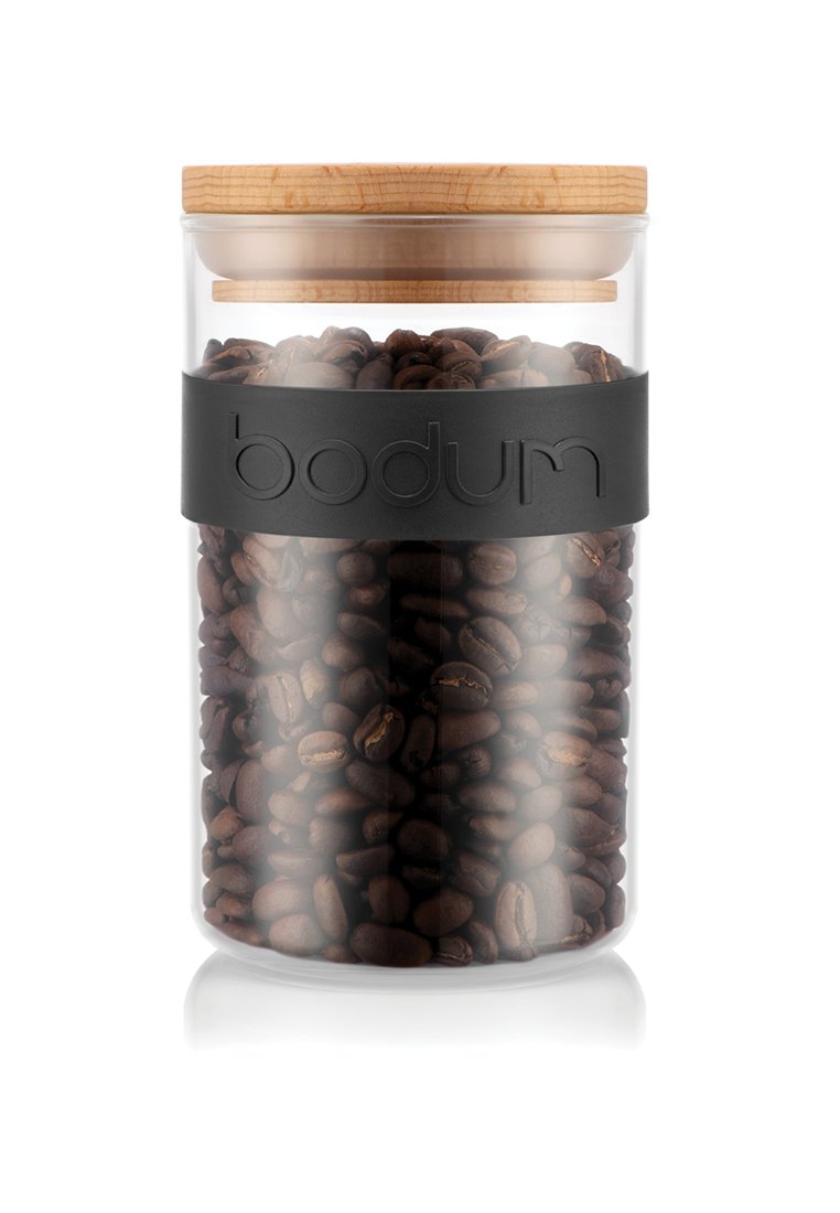 Tarro de cristal transparente con tapa de bambú, que contiene granos de café de color marrón oscuro. Presenta una banda de goma negra con el logotipo "bodum" en relieve.