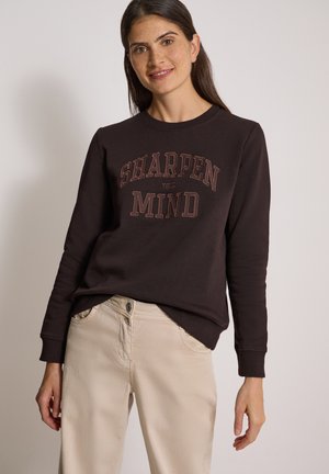 Frau mit langen dunklen Haaren lächelt, trägt einen schwarzen Sweatshirt mit dem Text "Sharpen Your Mind" und hellbeige Hosen vor einfarbigem Hintergrund.