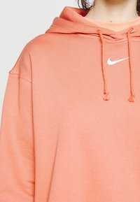 Oversized huvtröja i mjuk korallfärg, med en vit Nike-logotyp, justerbara dragsko och avslappnad passform.