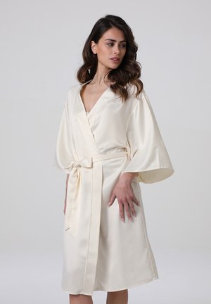 Donna con capelli castano ondulati che indossa un vestaglia di raso avorio lunghezza al ginocchio, legata in vita, in piedi davanti a uno sfondo grigio chiaro uniforme.