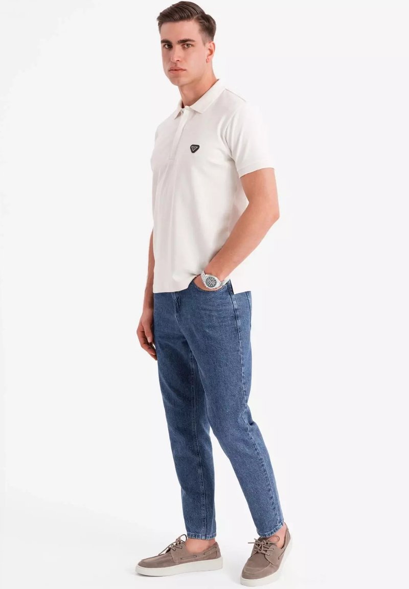 Hvid polo-shirt med et sort logo, parret med lyseblå tapered jeans og grå sko, stående mod en neutral baggrund.