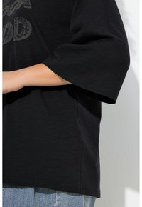 Zwart T-shirt met korte mouwen en een losse pasvorm, gemaakt van een gestructureerde stof met een subtiel grafisch ontwerp aan de voorkant. Gedragen over lichtblauwe spijkerbroek.