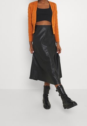 Osoba ubrana w pomarańczowy sweter na drutach, czarny krótki top, czarną skórzaną spódnicę midi oraz czarne sznurowane botki z klamrami, stojąca na tle białej ściany.