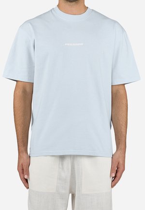 Mężczyzna ubrany w jasnoniebieską, oversize'ową koszulkę z napisem "PEGADOR" na środku klatki piersiowej oraz luźne białe spodnie.