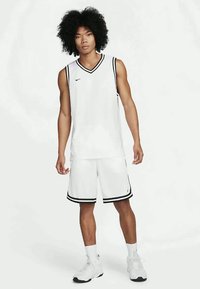 Hombre joven con cabello rizado, vestido con un uniforme de baloncesto blanco con ribetes negros, de pie con los brazos relajados contra un fondo blanco liso.
