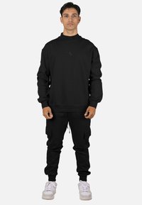 Sudadera negra con cuello alto y logo sutil, combinada con pantalones jogger negros con bolsillos laterales. Calzado: zapatillas blancas.