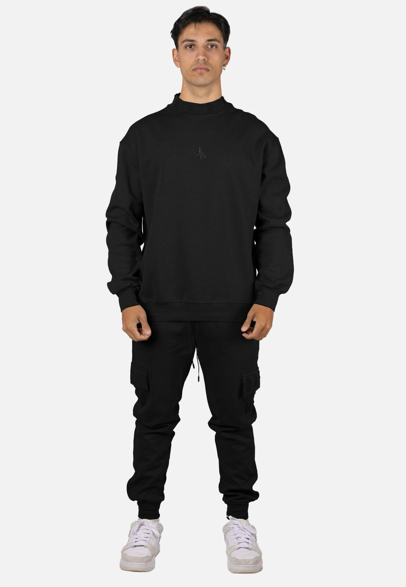 Sudadera negra con cuello alto y logo sutil, combinada con pantalones jogger negros con bolsillos laterales. Calzado: zapatillas blancas.