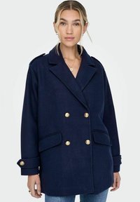 Manteau en laine bleu marine à double boutonnage avec des boutons dorés, revers pointus et poches latérales. Présente une coupe décontractée et des manches retroussées.