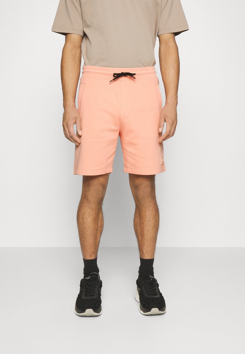 Shorts de sport couleur pêche avec une taille élastique, cordon de serrage noir, poches latérales et écusson logo au bas.