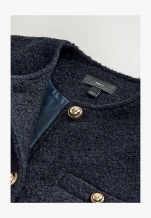 Námornícky textúrovaný kabát s mäkkým vlnitým povrchom, zlatým zapínaním na gombík a hladkým vnútorným podšívkou. Štítok uvádza "NEXT," vyrobené v Bangladéši.