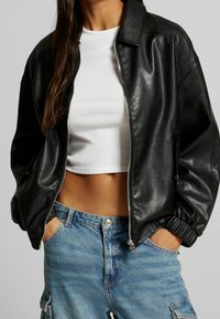 Svart fusk läder bomberjacka med dragkedja, ribbade ärmavslut och en framträdande krage. Matchas med en vit crop top och blå jeansshorts.