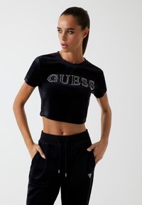 Juodas trumpas marškinėliai su "GUESS" blizgučiais, derinami su juodais aksominiais joggingais, turinčiais šonines kišenes ir mažą trikampį logotipo dizainą.