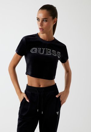 Zwarte cropped t-shirt met "GUESS" in rhinestones, gecombineerd met zwarte fluwelen joggers met zijzakken en een klein driehoekig logo ontwerp.