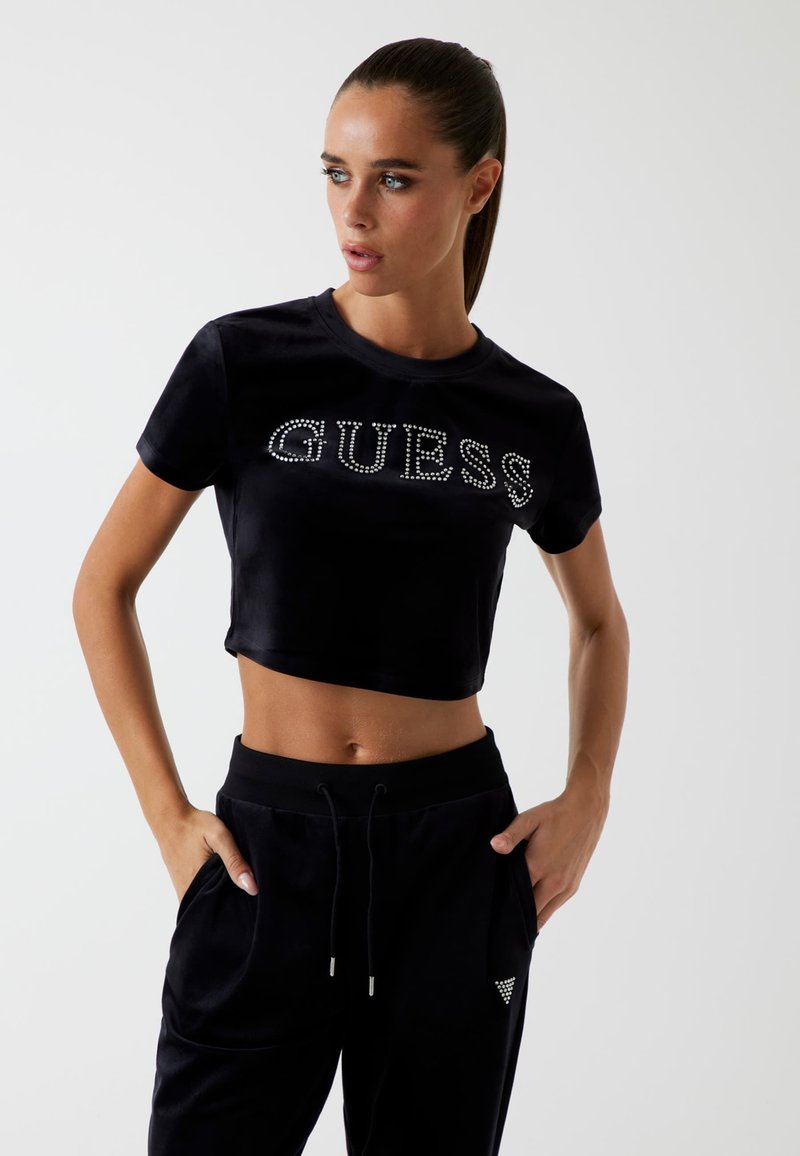 Black cropped t-shirt with "GUESS" in rhinestones, paired with black velvet joggers featuring side pockets and a small triangular logo design.