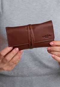 Brown leather wallet with textured surface, wrapped with a thin strap, featuring visible stitching and embossed branding detail.