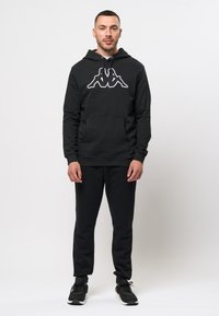 Zwarte hoodie met wit Kappa-logo en voorkraag, gecombineerd met zwarte broek en zwarte sportschoenen. Eenvoudig, comfortabel ontwerp.