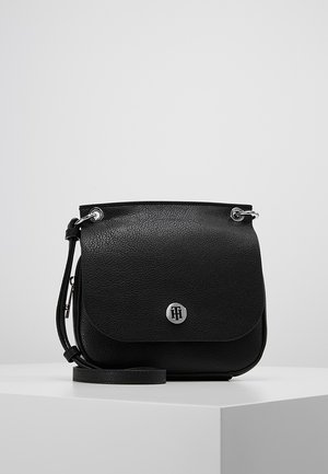 Schwarze, strukturiert genarbte Leder-Umhängetasche mit Klappe, silbernem Logo-Emblem und verstellbarem Riemen, präsentiert auf einem weißen Podest.