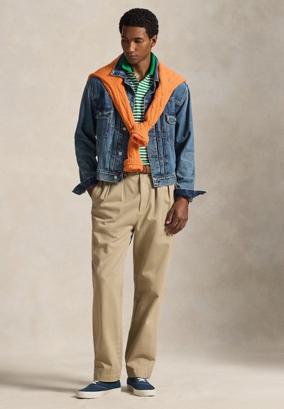 Άνδρας που φοράει πράσινη ριγωτή πόλο μπλούζα, μπλε τζιν μπουφάν, πορτοκαλί πουλόβερ περασμένο στους ώμους, μπεζ παντελόνι και μπλε αθλητικά παπούτσια στέκεται μπροστά σε ουδέτερο φόντο.