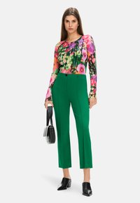 Bluse mit langen Ärmeln in floralen Mustern und lebhaften Farben; grüne taillierte Hose mit geradem Schnitt; schwarze Handtasche und Schuhe mit Blockabsatz vervollständigen den Look.