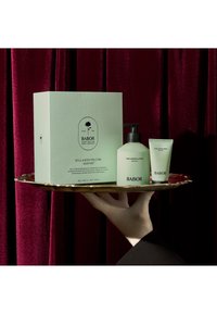 Babor huidverzorgingsset op een gouden dienblad: mintgroene textuurdoos, lotion met zwarte pomp en kleine wasbuis, tegen een wijnrode achtergrond.